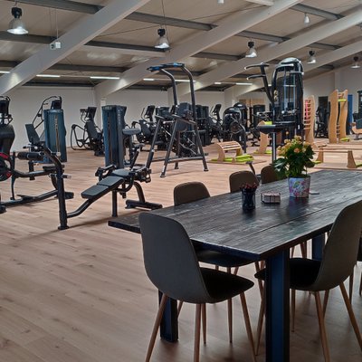 Großes Fitnessstudio mit vielen Trainingsgeräten im Hintergrund sowie einem Tisch mit Stühlen im Vordergrund.